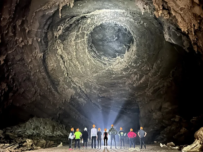 Nhóm khách trekking đội đèn pin đứng dưới một khối đá tròn khổng lồ trong hang Phong Nha.