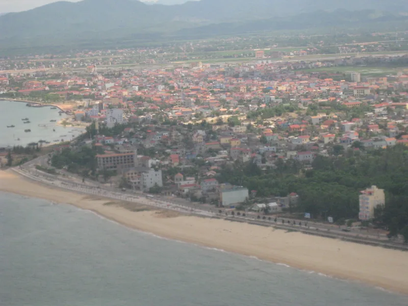Toàn cảnh trên không thành phố Đồng Hới với bãi biển cát, đường ven biển và nhà cửa dày đặc phía sau là núi.