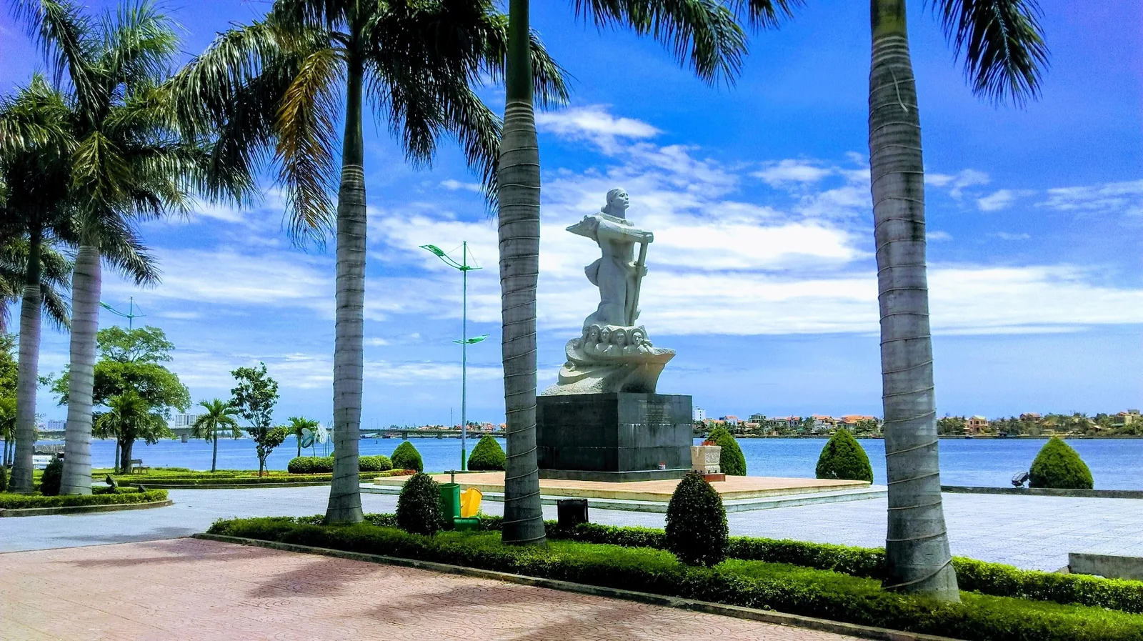 Bức tượng đá đứng trên quảng trường bên sông, rợp bóng cây dừa dưới trời xanh ở Đồng Hới.