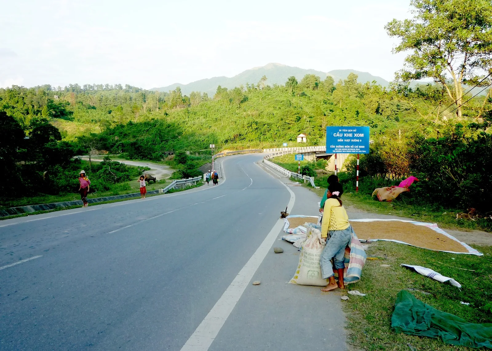 Con đường nông thôn xuyên đồi núi xanh gần điểm vượt đường 9 của đường mòn Hồ Chí Minh ở Quảng Trị, người dân phơi lúa bên.
