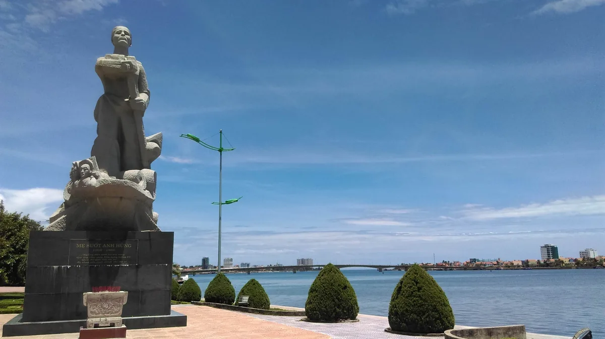Tượng đá trên bệ đứng nhìn ra sông Nhật Lệ gần Đồng Hới, phía xa có cầu và đường chân trời thành phố.