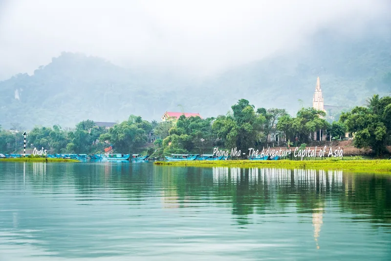 Núi mờ sương phía sau ngôi làng ven sông với tháp chuông nhà thờ ở Phong Nha, thuyền neo đậu trên mặt nước yên bình.