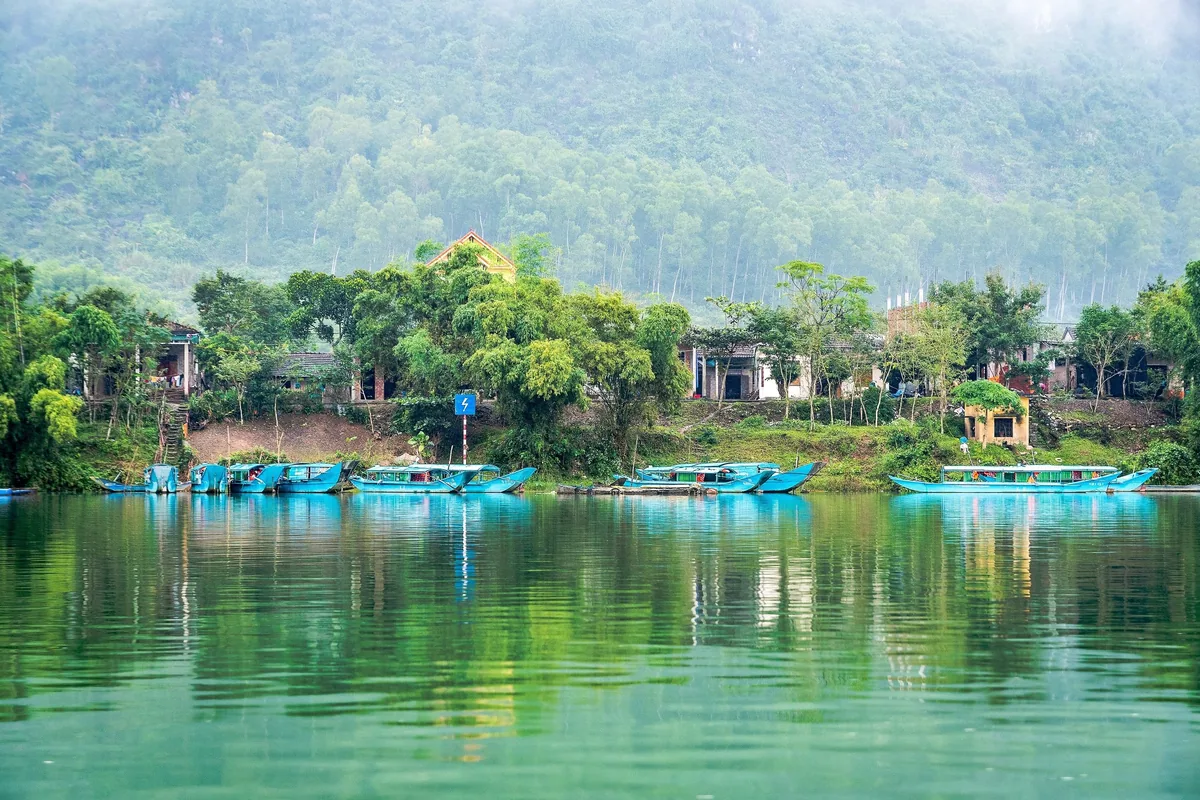 Những chiếc thuyền du lịch màu xanh đậu bên bờ sông Phong Nha, phía sau là núi đá vôi mờ sương và ngôi làng nhỏ.